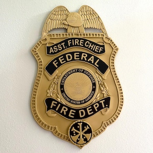 May include: A gold-colored metal badge with the words "ASST. FIRE CHIEF FEDERAL FIRE DEPT." and a seal with the words "DEPARTMENT OF DEFENSE UNITED STATES OF AMERICA".