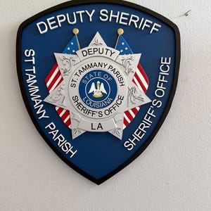 May include: A blue and white badge with the text "DEPUTY SHERIFF ST. TAMMANY PARISH SHERIFF'S OFFICE LA" and a star with an American flag on each point.