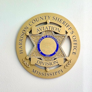 May include: A gold-colored badge with the words "Harrison County Sheriff's Office Aviation Division Mississippi" around the edge. The center of the badge features a five-pointed star with the words "State of Mississippi" and "1841" inside.