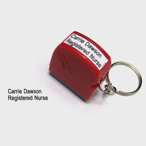May include: A red stamp with the text "Carrie Dawson Registered Nurse" printed on a white label. The stamp is attached to a silver keyring. The text "Carrie Dawson Registered Nurse" is also printed on the side of the stamp.