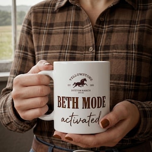以下が含まれることがあります： 「Yellowstone Dutton Ranch」と「Beth Mode activated」の文字が茶色で書かれた白いセラミックマグ。マグには馬のロゴがあり、茶色のチェック柄のシャツを着た人が持っています。