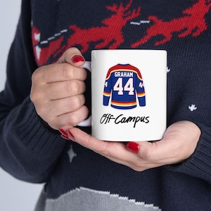 Puede incluir: Taza de cerámica blanca con un diseño de camiseta de hockey azul, roja y amarilla, que muestra el nombre "Graham" y el número "44". La taza también tiene el texto "Off-Campus" en una fuente manuscrita.