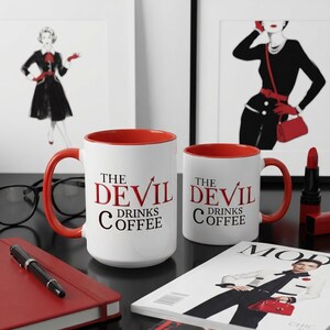 May include: Two white coffee mugs with red interiors and handles. The mugs have the text "THE DEVIL DRINKS COFFEE" in red and black. The mugs are on a black surface with a red notebook, pen, and magazine.