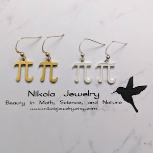 Puede incluir: Dos pares de pendientes con el símbolo matemático pi. Un par es dorado y el otro es plateado. Los pendientes cuelgan de una superficie blanca.