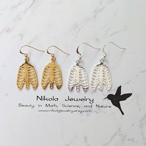 May include: Two pairs of earrings, one gold and one silver, featuring a ribcage design. The earrings are hanging from a white surface. The text "Nikola Jewelry" is visible below the earrings.