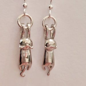 May include: Silver dangle earrings featuring a small, detailed cat charm hanging upside down.