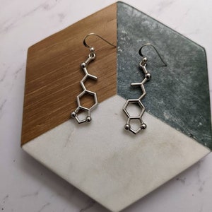 May include: A pair of silver earrings shaped like the chemical structure of dopamine. The earrings are hanging from a wooden and marble display stand.