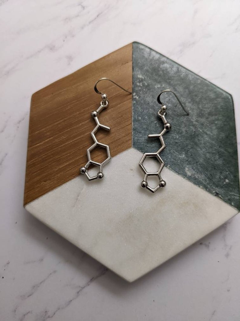 May include: A pair of silver earrings shaped like the chemical structure of dopamine. The earrings are hanging from a wooden and marble display stand.