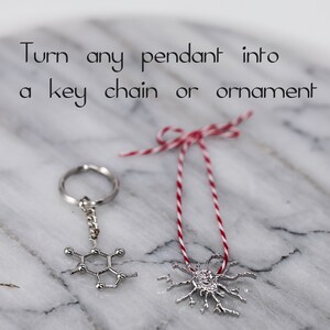 May include: Two silver pendants, one shaped like a molecule and the other like a snowflake, are shown on a white marble surface. One pendant is attached to a silver key chain and the other is attached to a red and white string.