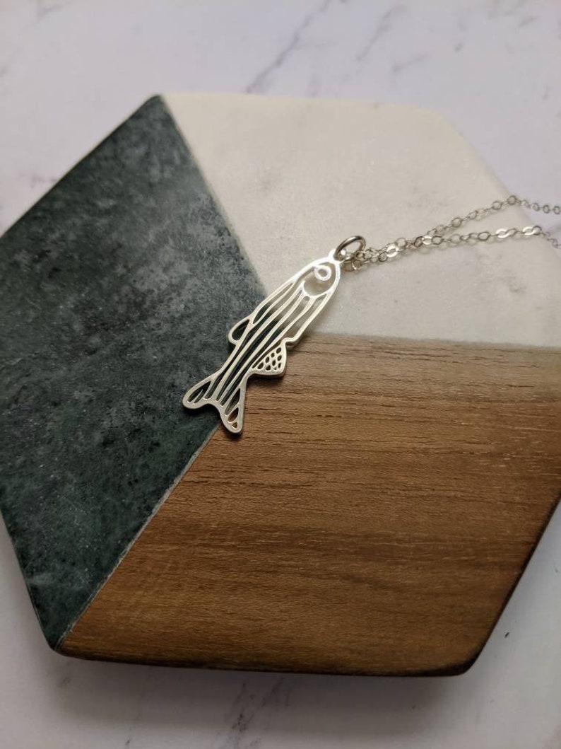 May include: A silver necklace with a detailed fish pendant. The pendant is cut out and has a delicate design. The necklace is on a wooden and green marble surface.