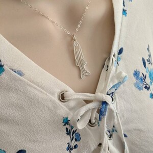 May include: A silver necklace with a fish pendant. The fish is detailed with lines and has a slightly curved shape. The necklace is on a model wearing a white blouse with blue floral print.