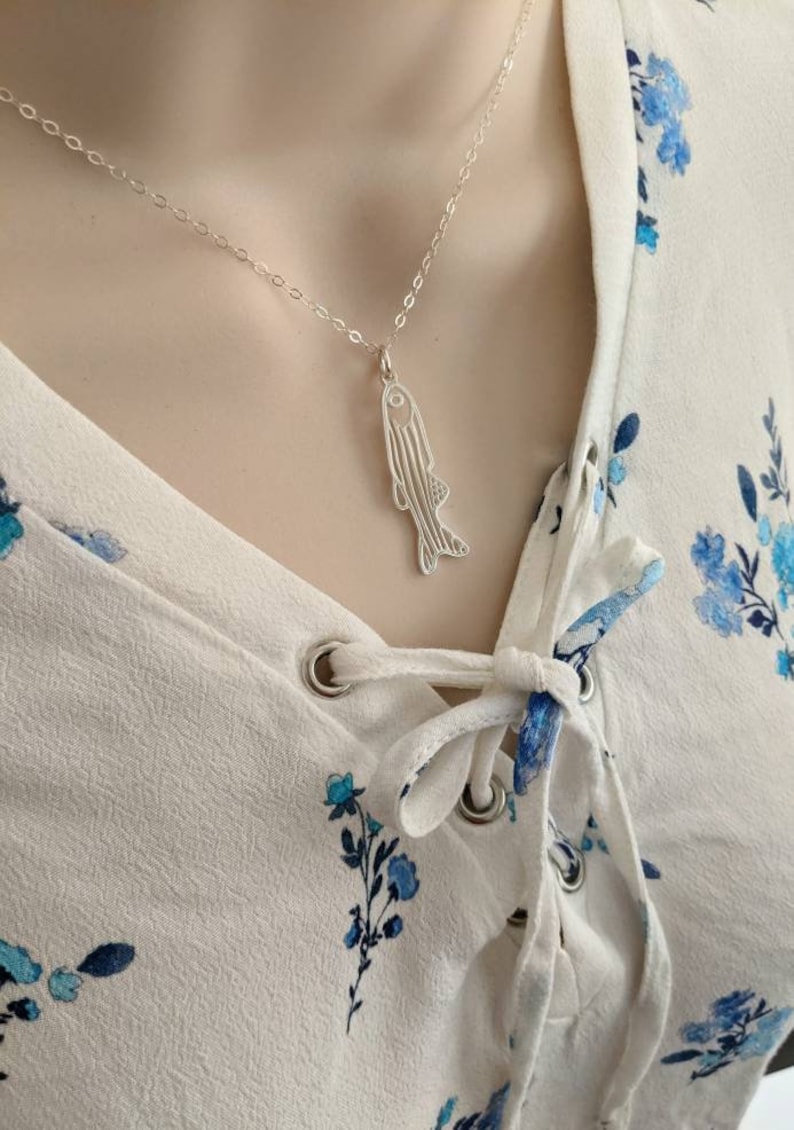 May include: A silver necklace with a fish pendant. The fish is detailed with lines and has a slightly curved shape. The necklace is on a model wearing a white blouse with blue floral print.