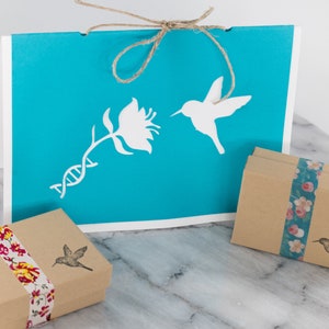 May include: A turquoise blue card with a white hummingbird, flower, and DNA strand design. The card is tied with twine and comes with two small brown boxes wrapped in floral patterned paper.