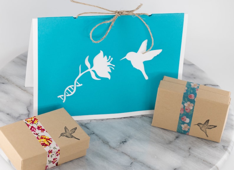 May include: A turquoise blue card with a white hummingbird, flower, and DNA strand design. The card is tied with twine and comes with two small brown boxes wrapped in floral patterned paper.