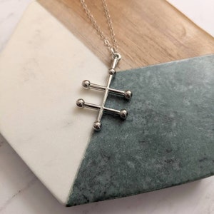 May include: A silver necklace with a geometric pendant featuring a stylized tree design with five round balls. The necklace is displayed on a wooden and marble surface.