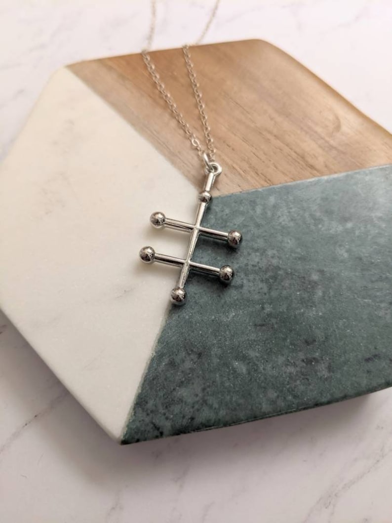 May include: A silver necklace with a geometric pendant featuring a stylized tree design with five round balls. The necklace is displayed on a wooden and marble surface.