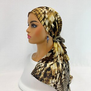 May include: A brown and gold patterned headscarf with a snake skin design. The scarf is tied in a knot at the back of the neck and the ends are draped down the shoulders.
