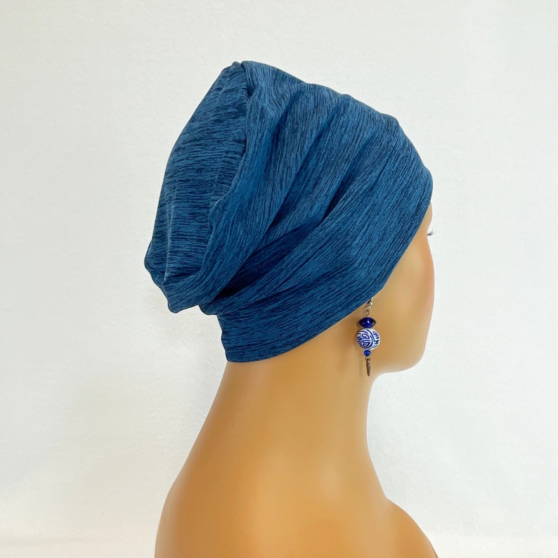 May include: A blue knit beanie hat with a textured appearance. The hat is worn on a mannequin head, paired with blue and white earrings. The earrings have a floral design and are suspended from a silver hook.