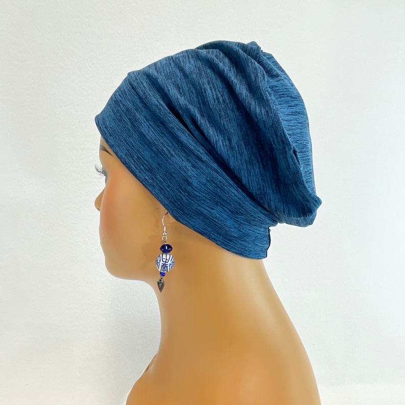May include: A blue beanie-style hat with a textured pattern. The hat is worn with a pair of dangling earrings. The earrings have blue and white beads and a silver-colored charm. The background is plain white.