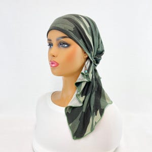 May include: A green and black camouflage head covering. The head wrap is tied at the back and has a flowing, draped design. The fabric has a pattern of various shades of green and black. The head covering is shown on a mannequin.