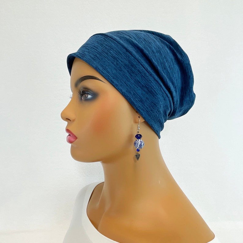 May include: A mannequin wearing a dark blue beanie and dangling earrings. The earrings feature blue and white ceramic beads with a geometric design, and a silver-colored charm. The beanie is made of a soft, textured fabric.