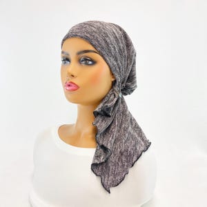 May include: Gray and black patterned head scarf with a ruffled edge. The scarf is tied in a knot at the back of the head and the ends are draped down the back.