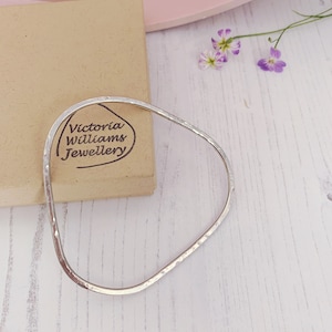 May include: A hammered silver-tone bracelet with an irregular, geometric shape. The bracelet is next to a brown box with the text "Victoria Williams Jewellery" and a few small purple flowers.