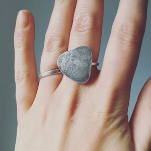 Könnte beinhalten: Ein silberner Ring mit einem herzförmigen Anhänger, der ein strukturiertes Blattmuster aufweist. Der Ring hat ein schmales Band. Der Ring wird an einem Finger getragen. Das Design des Anhängers erinnert an Baumzweige.