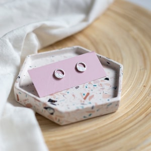 Könnte beinhalten: Silberne Ohrstecker mit strukturiertem Design in einem kreisförmigen Rahmen, präsentiert auf einer rosa Karte. Die Karte liegt auf einem sechseckigen Tablett mit Terrazzo-Muster. Das Tablett steht auf einer Holzoberfläche, mit weißem Stoff im Hintergrund.