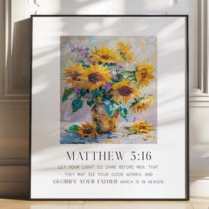 May include: A framed print featuring a vibrant watercolor painting of sunflowers in a vase. The artwork includes the text "MATTHEW 5:16" and a related Bible verse. The print is displayed on a wooden floor against a white wall.