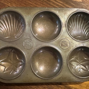 May include: Vintage six-cup muffin tin with decorative shell and leaf designs. The metal pan has a well-worn, aged appearance. The text "Minutes Made" and "Made in U.S. America" are visible.