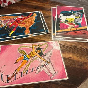 May include: Comic book art prints featuring DC Comics superheroes. The Flash is depicted running across a map of the United States. Batman and Robin are on a rooftop. Wonder Woman is shown with a lasso and a jet.