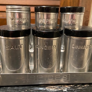 May include: A vintage metal spice rack with six silver canisters with black lids. The canisters are labeled "Salt", "Paprika", "Pepper", "Nutmeg", "Ginger", and "Cinnamon". The rack is labeled "SPICES".