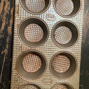 May include: A vintage silver-colored muffin tin with six individual cups. The tin has a textured surface and the words "EKCO" and "U.S.A. MADE" are printed on the bottom.