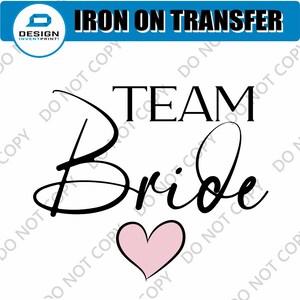 May include: Iron-on transfer design featuring the text "TEAM Bride" in black cursive font with a pink heart below.
