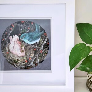 May include: Framed artwork featuring a blue bird tending a nest with a human heart inside. The heart is nestled in a woven nest with branches and red berries. The artwork is framed in a white square frame.