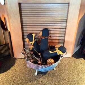 May include: Three black fabric birds with yellow ribbon accents are arranged in a metal colander with brown twigs and berries. The colander is sitting on a brown speckled countertop.