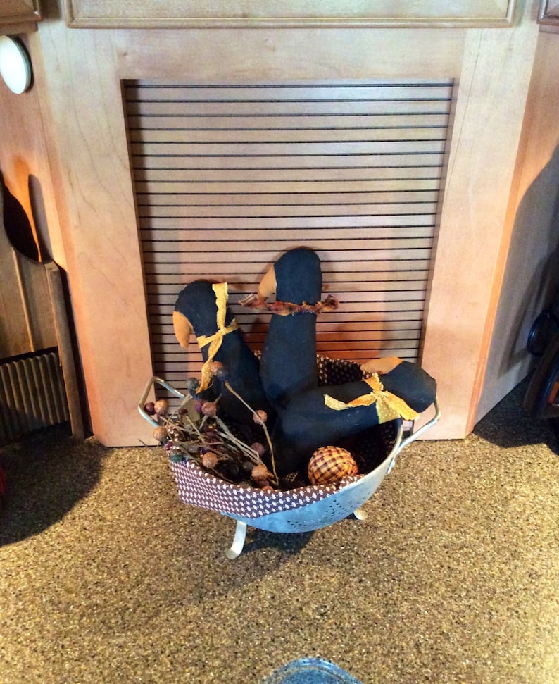May include: Three black fabric birds with yellow ribbon accents are arranged in a metal colander with brown twigs and berries. The colander is sitting on a brown speckled countertop.