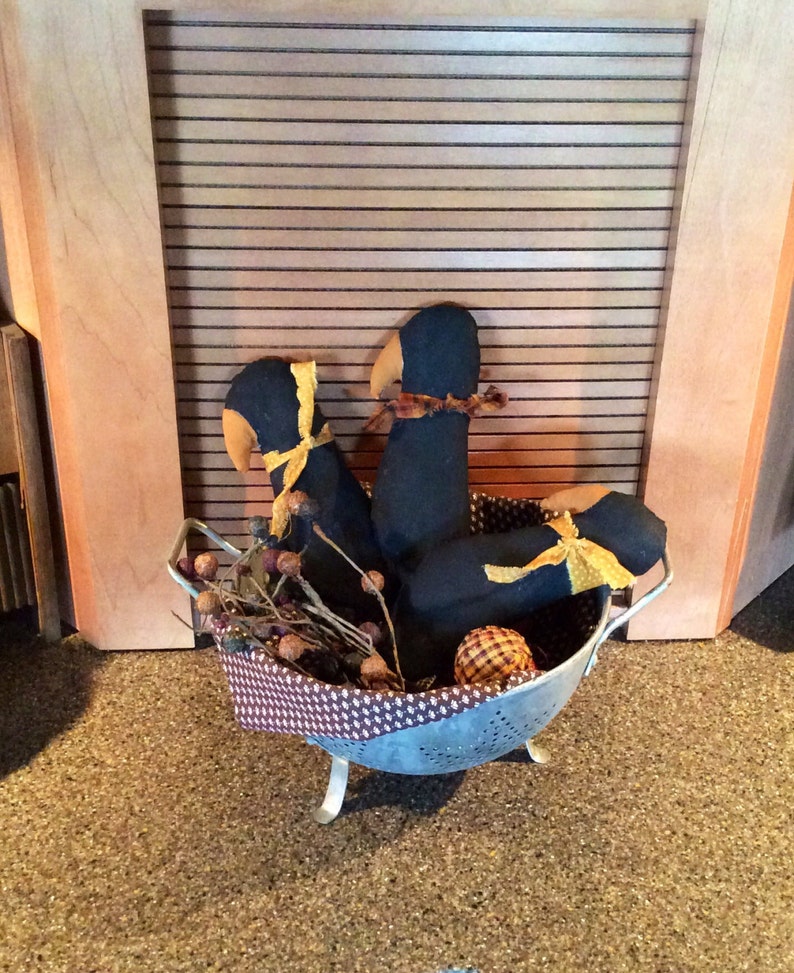May include: Three black fabric birds with yellow ribbon accents are arranged in a silver colander with brown twigs and acorns. The colander is sitting on a brown carpet.