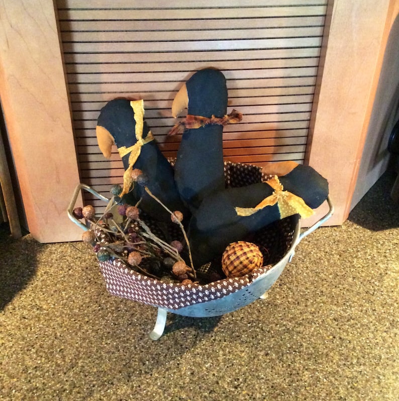 May include: Three black fabric birds with yellow ribbon accents are arranged in a metal colander with brown and white checkered lining. The colander is on a brown surface.
