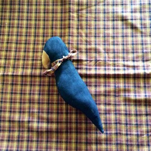May include: A black fabric bird with a brown fabric beak and a yellow and brown fabric bow tied around its neck. The bird is lying on a plaid fabric surface.