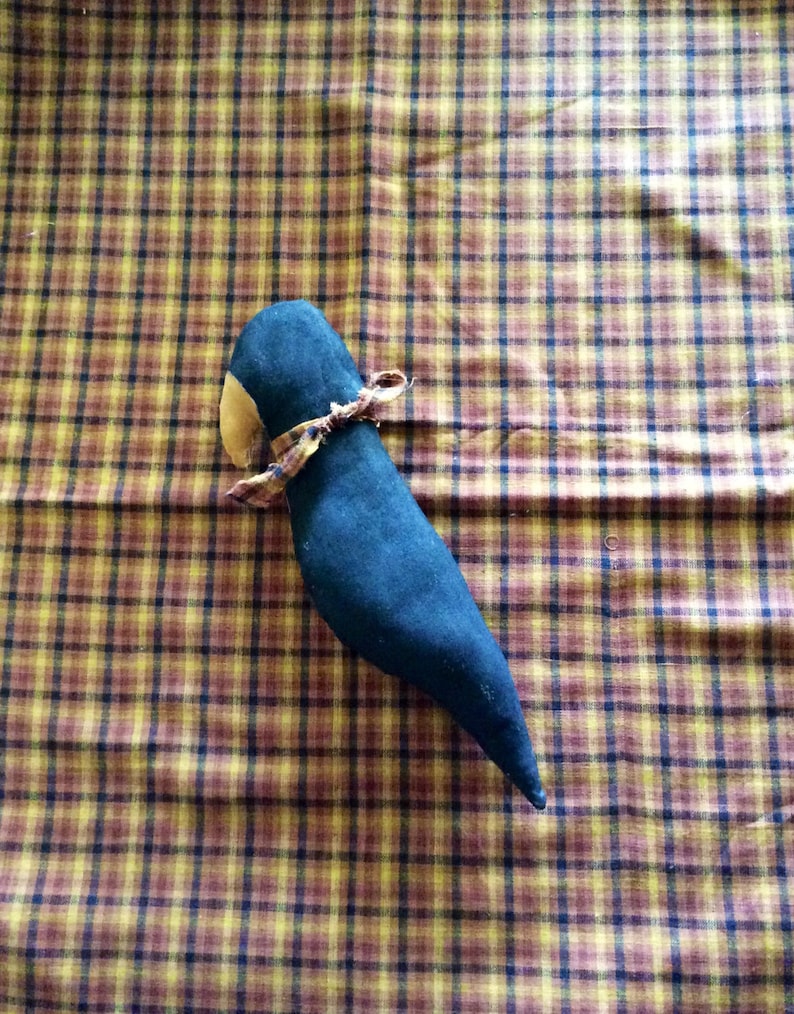 May include: A black fabric bird with a brown fabric beak and a yellow and brown fabric bow tied around its neck. The bird is lying on a plaid fabric surface.
