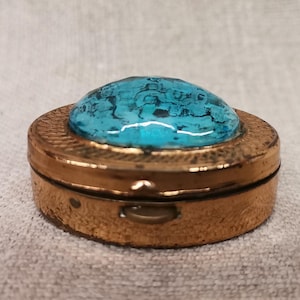 May include: A small, round, antique-style pillbox with a gold-coloured metal base. The lid features a vibrant turquoise stone cabochon. The box is closed, and the surface has a textured appearance. The pillbox is resting on a neutral-coloured fabric.