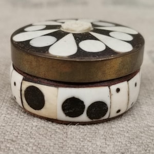 May include: A small, round decorative box with a brass rim. The lid features a black and white floral pattern, while the body is adorned with black dots and white rectangular shapes. The box is likely used for storing small items.