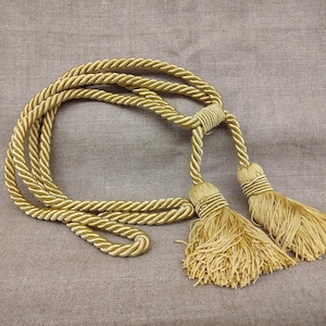 Pode incluir: Um cordão decorativo de cor dourada com um design de corda torcida e duas borlas a condizer nas extremidades. O cordão está enrolado e preso com um nó central, exibindo um brilho metálico e franjas finas e densas em cada borla.