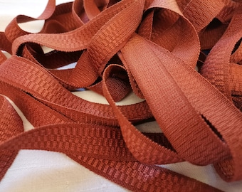 May include: A close-up of a pile of rust-colored fabric trim with a textured pattern. The trim is made of a soft, woven material and has a subtle sheen.