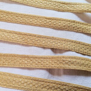 May include: Close-up of several strands of woven, golden-coloured ribbon. The ribbon has a textured, patterned surface and is arranged diagonally across a white background. The ribbon appears to be made of a natural fibre.