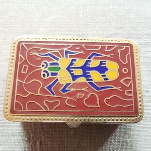 May include: A small, rectangular, gold-colored box with a red, blue, yellow, and green enamel design of a stylized insect. The box has a decorative border of gold dots.