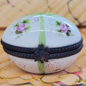 May include: A white porcelain egg-shaped trinket box with a hinged lid. The box is decorated with hand-painted pink roses and green leaves. The lid has a metal trim and a decorative metal clasp.