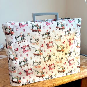 May include: A rectangular sewing machine cover featuring a white fabric base with a repeating pattern of vintage-style sewing machines in shades of green and pink, accented by small floral motifs and spools of thread, with a grey plastic handle.